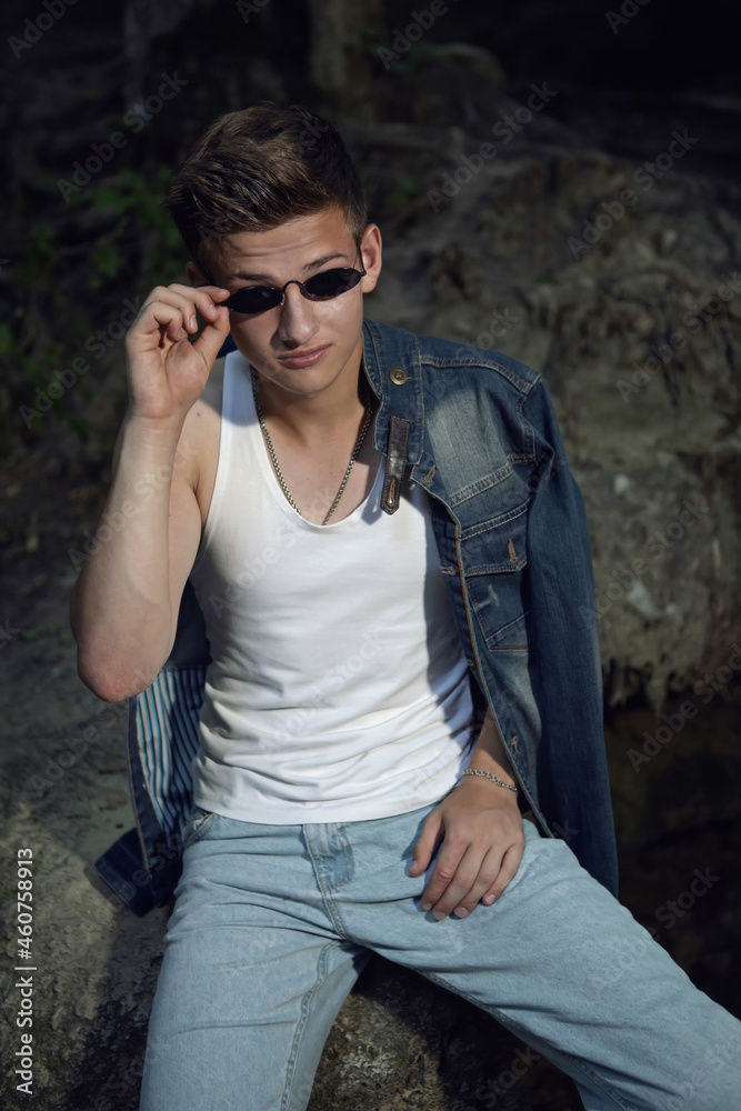 sexy trendy teen guy in a white T-shirt and blue jeans is sitting on a rock by a waterfall
