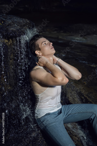sexy trendy teen guy in a white T-shirt and blue jeans is sitting on a rock by a waterfall