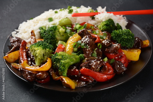 Asian teriyaki beef with red and yellow bell peppers, broccoli and sesame seeds on a plate on the table. Spicy teriyaki beef stir fry with vegetables and rice on a dark background