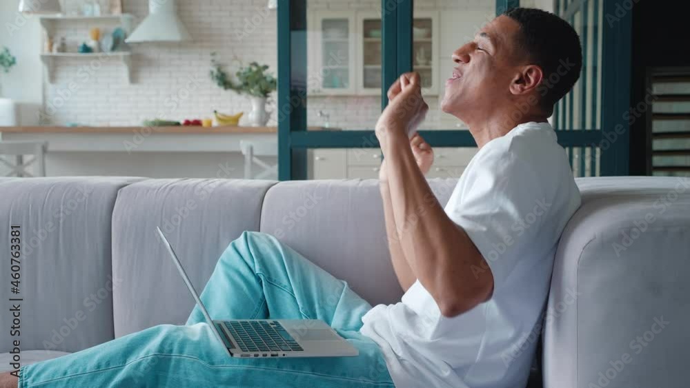 Handsome African American man winner resting on sofa rejoicing looking at laptop monitor, dancing, clenching fists after receiving good news. Male celebrating success bet win at home on sofa Handsome African American man winner resting on sofa rejoicing looking at laptop monitor, dancing, clenching fists after receiving good news. Male celebrating success bet win at home on sofa