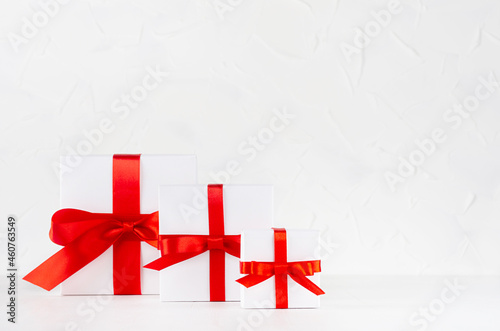 White present boxes different size with red satin ribbon and bow on white table and soft light wall, copy space.