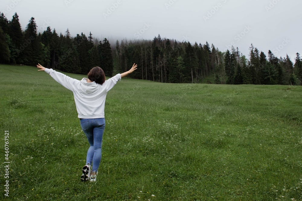 Woman traveler standing on a glade high in the mountains in front of forest, space for text, atmospheric epic moment