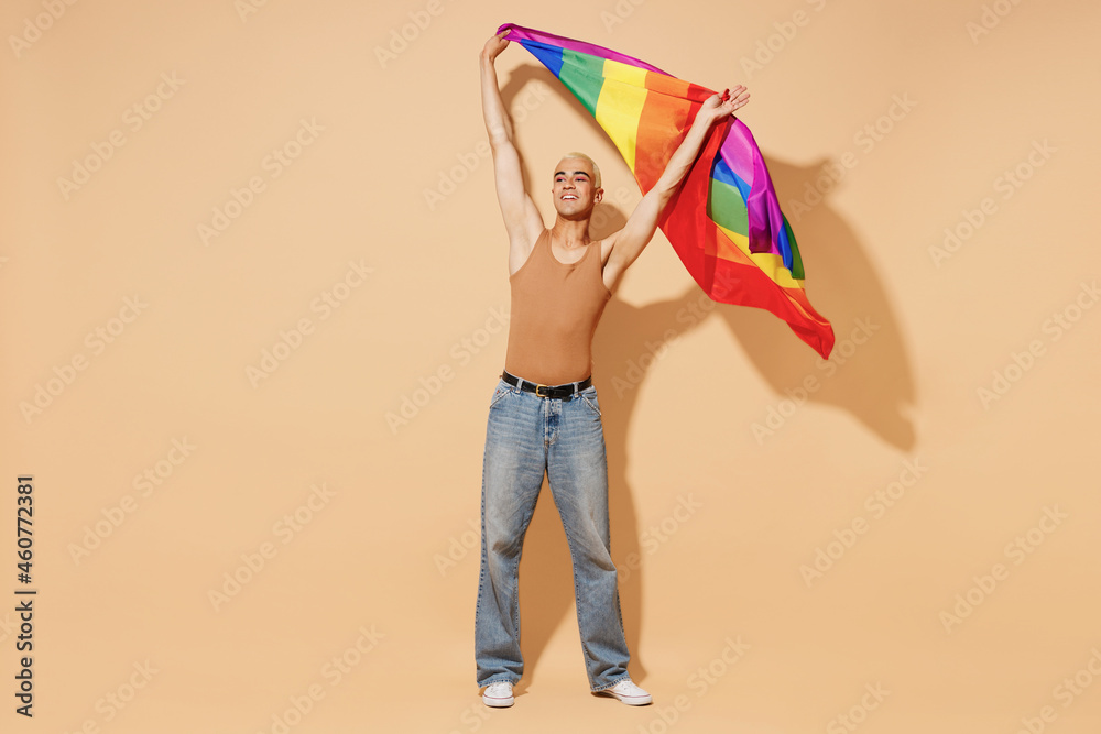 Full body young activist bisexual happy fun blond latin gay man with make up in beige tank shirt waving hold rainbow flag isolated on plain light ocher background studio People lgbt lifestyle concept - obrazy, fototapety, plakaty 