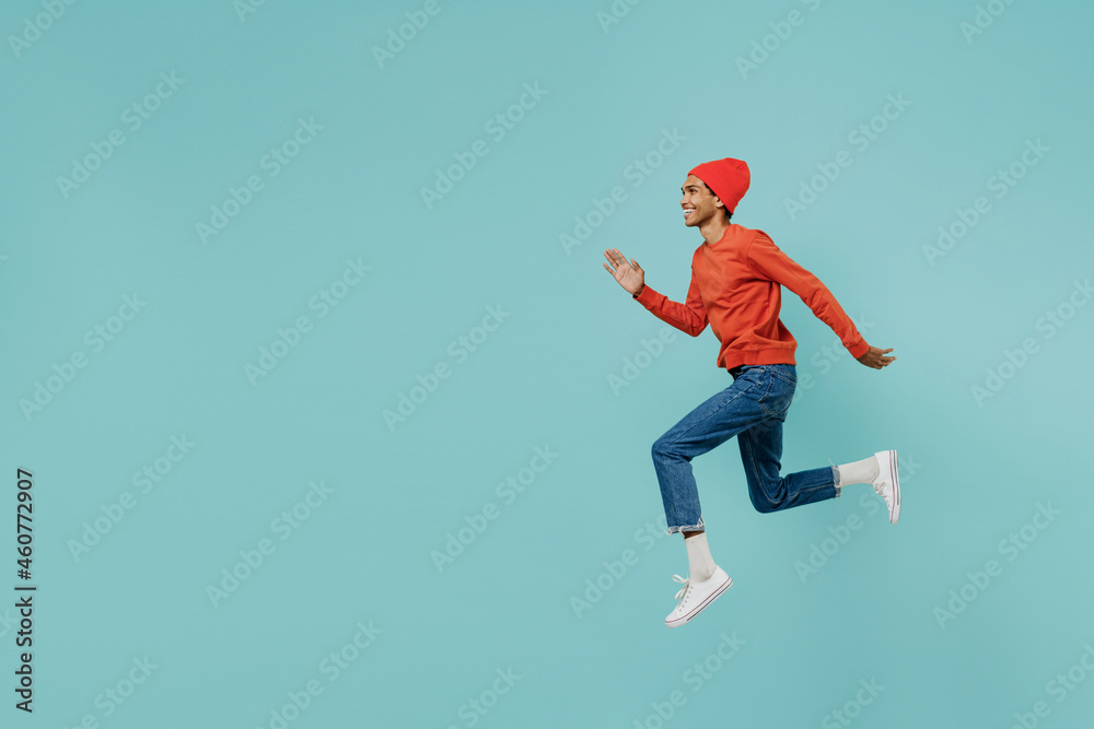 Full body side view fun cool young smiling happy african american man in orange shirt hat jump high run fast isolated on plain pastel light blue background studio portrait People lifestyle concept.