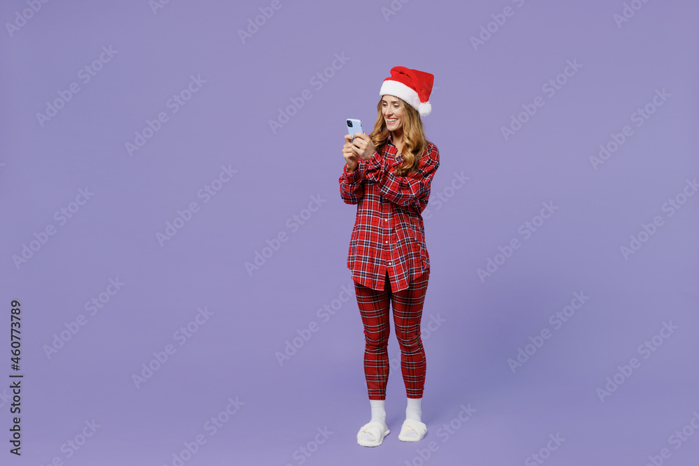 Obraz premium Full size body length young Santa woman 30s wears shirt Christmas hat hold in hand use mobile cell phone isolated on pastel light violet background studio portrait. Happy New Year celebration concept.