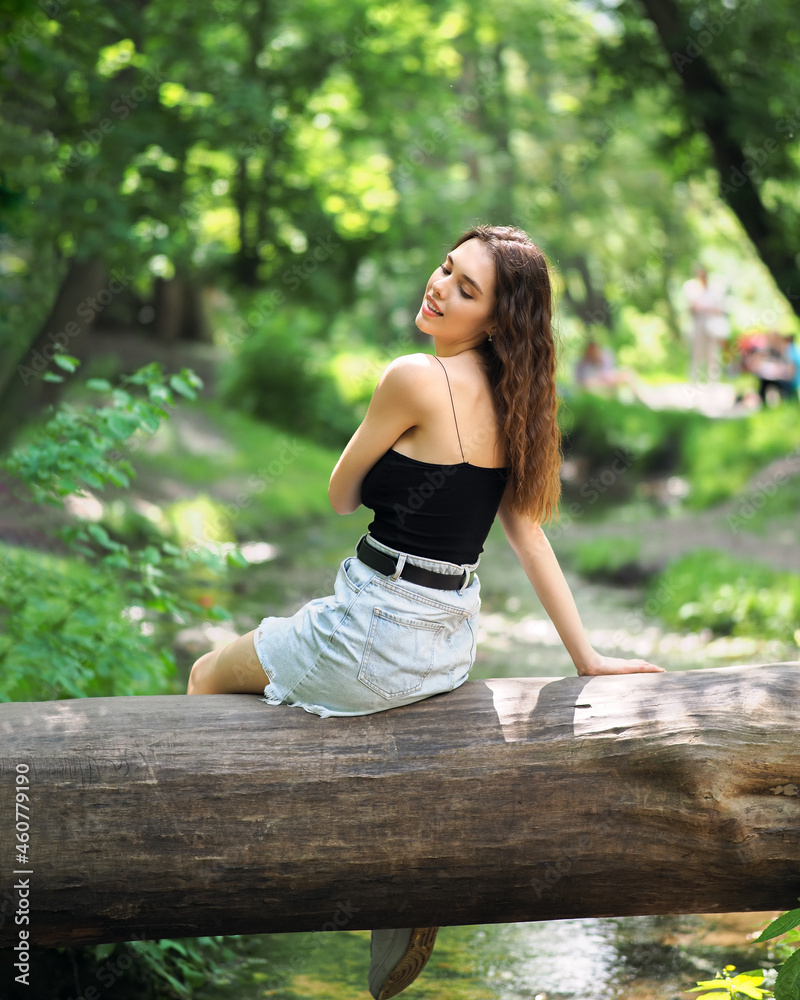 Naklejka premium A woman in a denim skirt and a top is sitting on a log in the forest