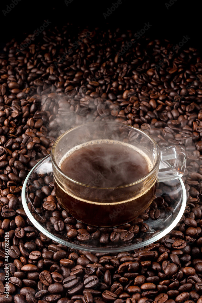 Fototapeta premium Glass of hot black coffee with glass saucer place on pile of coffee seed. hot dark coffee and vapour. close up. top view.