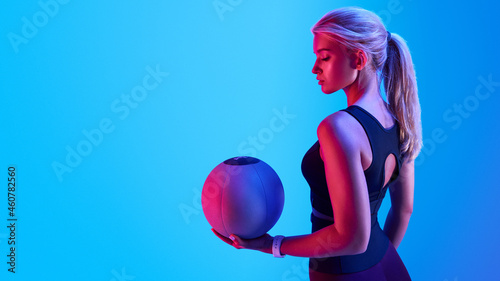 Attractive blonde sportswoman portrait holding with medicine fitness ball neon style creative light. Confident fitness woman standing with a medicine ball