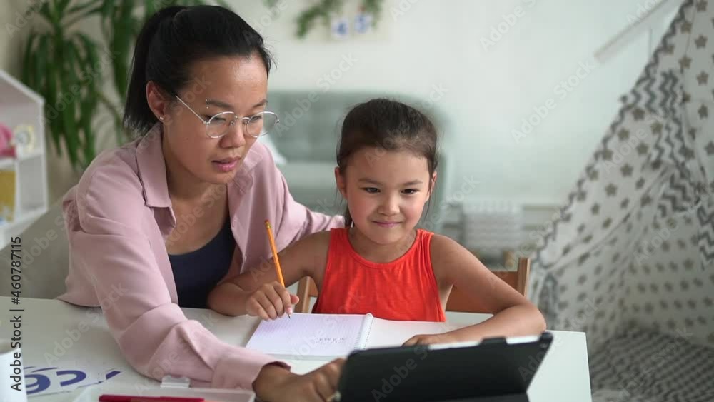 Online primary school family education. Spbd Concentrated Korean mom with glasses shows little daughter information on tablet at table in light room Online primary school family education. Spbd Concentrated Korean mom with glasses shows little daughter information on tablet at table in light room
