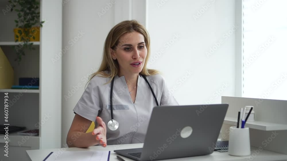 Female doctor talking and smiling in front of laptop during online consultation in clinic spbd. Beautiful young woman looks at computer screen and speaks with smile, sits at desk in light interior Female doctor talking and smiling in front of laptop during online consultation in clinic spbd. Beautiful young woman looks at computer screen and speaks with smile, sits at desk in light interior