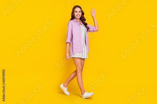 Full length photo of happy positive woman walk empty space wave hand hello good mood isolated on yellow color background