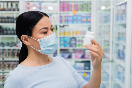 asian woman in medical mask looking at bottle with vitamins in drugstore