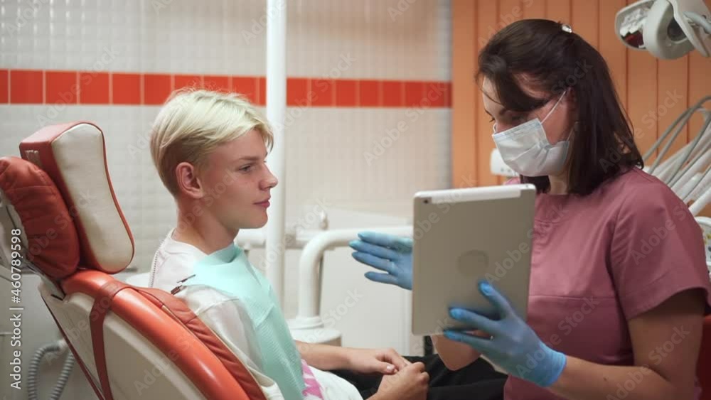 Woman orthodontist talking with patient and using tablet while sitting in clinic office spbd. Close-up view of young female doctor talks to teenager and shows result on screen of device, he looks with