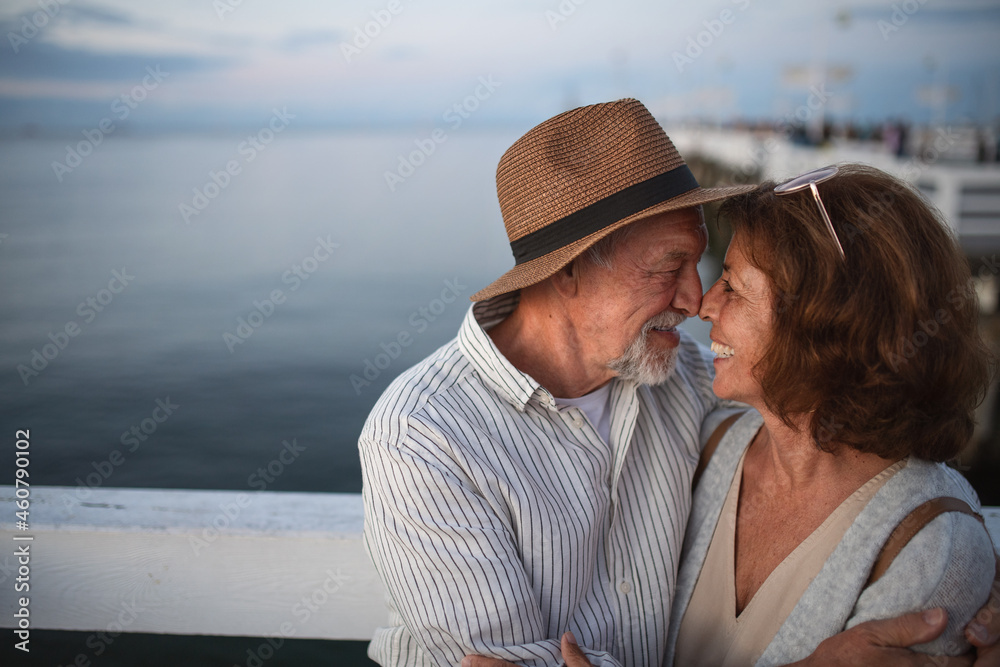 Portrait of happy senior couple in love hugging outdoors on pier by sea, looking at each other, summer holiday.