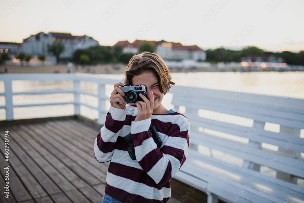 Preteen girl tourist taking photo with camera on pier by sea at sunset, summer holiday concept.
