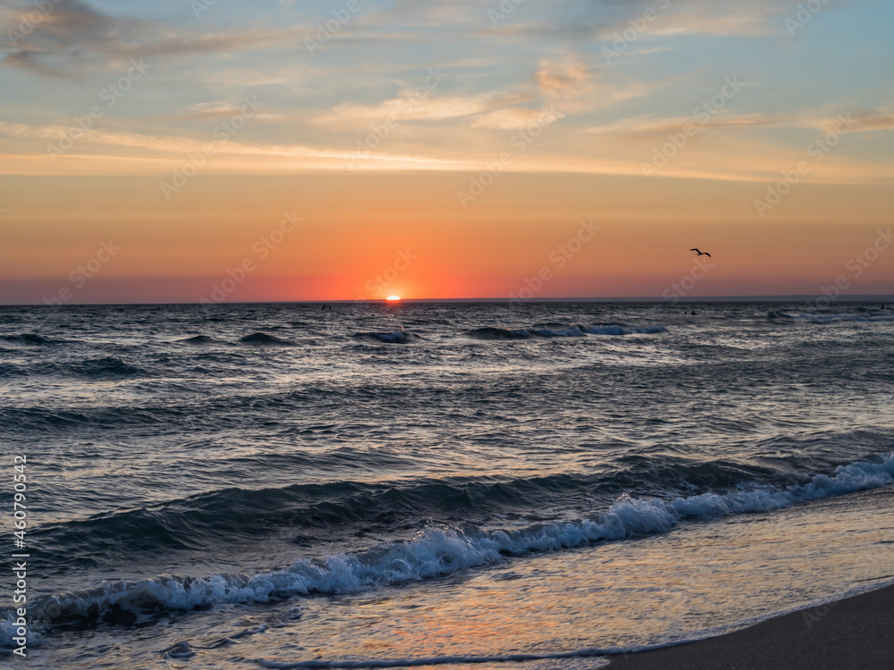 Fototapeta premium Picturesque landscape. Sunset over the sea. There are clouds in the sky. Silhouettes of a seagull flying over the sea. Sandy coastline. Beautiful big waves. Horizontally