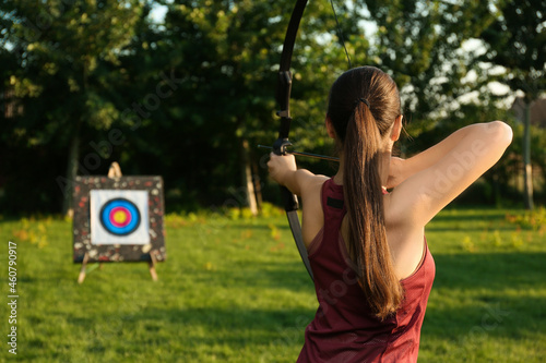 Slika na platnu Woman with bow and arrow aiming at archery target in park, back view