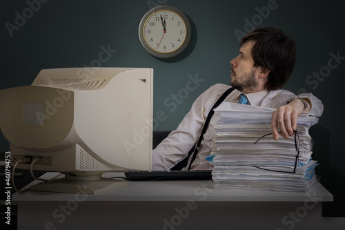 Tired man is looking on clock in office at night. Overwork concept. photo