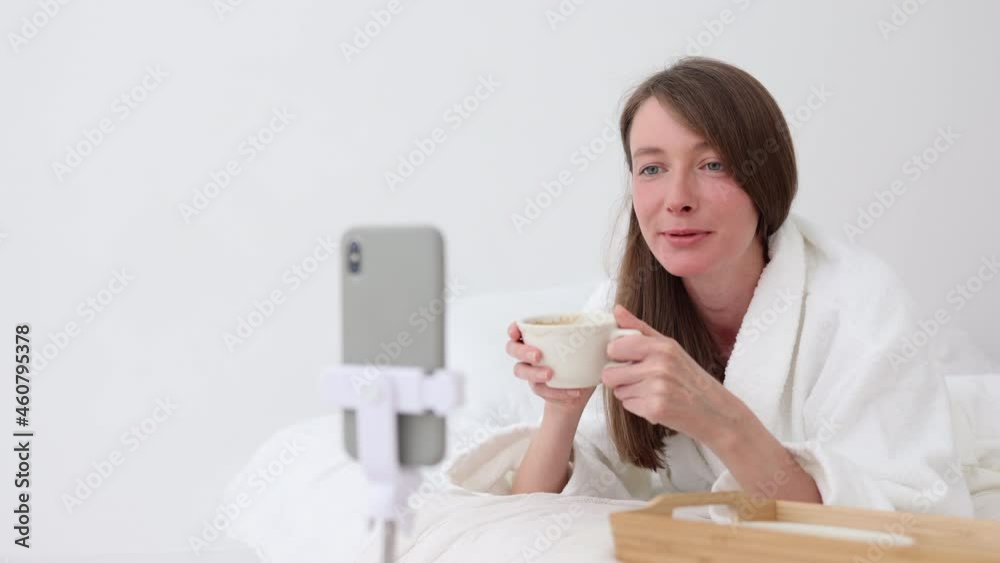 Woman talking in front of smartphone and drinking coffee, lying on bed in home bedroom spbi. Close view of young beautiful female waves hand in front of phone screen and talks with smile, holds cup in Woman talking in front of smartphone and drinking coffee, lying on bed in home bedroom spbi. Close view of young beautiful female waves hand in front of phone screen and talks with smile, holds cup in