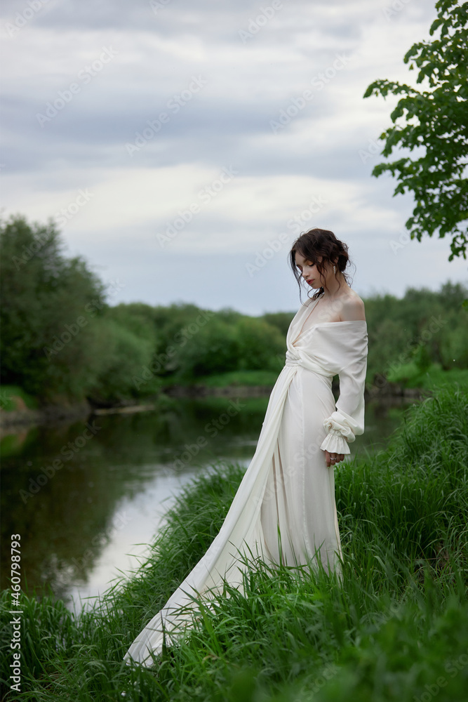 Beautiful young woman in white dress resting on shore lake pond river. Professional makeup and hairstyle with curled hair Beautiful young woman in white dress resting on shore lake pond river. Professional makeup and hairstyle with curled hair