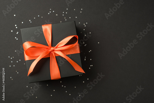 Top view photo of stylish black giftbox with orange satin ribbon bow and bright sequins on isolated black background with copyspace