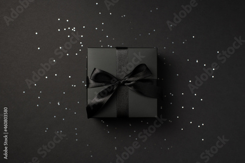 Top view photo of black giftbox with black satin ribbon bow over shiny sequins on isolated black background
