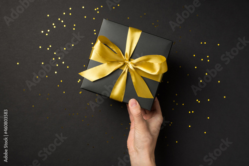 First person top view photo of hand holding black giftbox with yellow satin ribbon bow over shiny golden sequins on isolated black background with empty space
