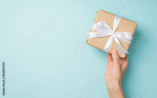 First person top view photo of hand holding stylish craft paper giftbox with white satin ribbon bow on isolated pastel blue background with copyspace