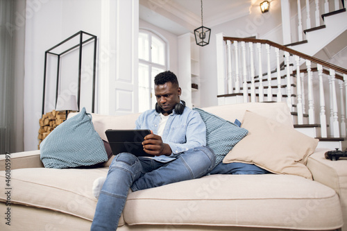 Young african man in wireless headphones resting on comfy couch with digital tablet in hands. Handsome guy in casual clothes surfing internet during free time at home.