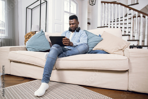 Handsome young African American guy in denim shirt and jeans sitting on comfy sofa and using digital tablet with wireless headphones. Concept of people, technology and lifestyles.