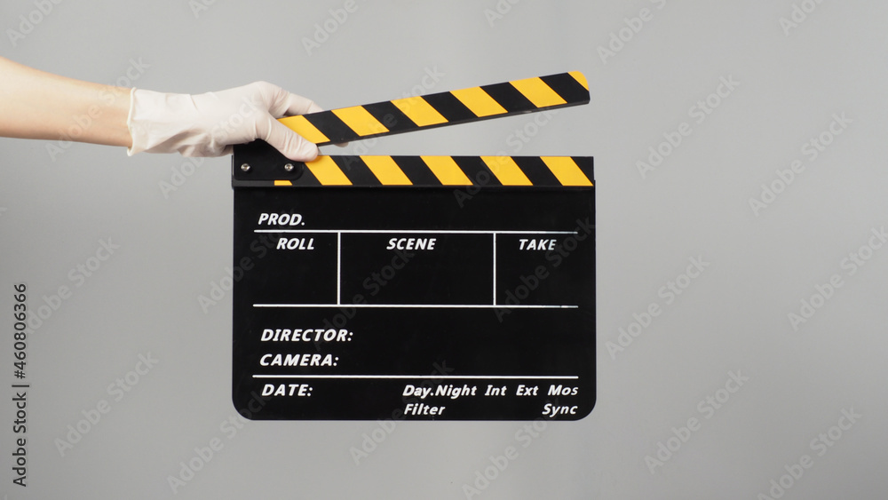 Hand is holding yellow with black clapper board color and wear white medical glove on grey background.