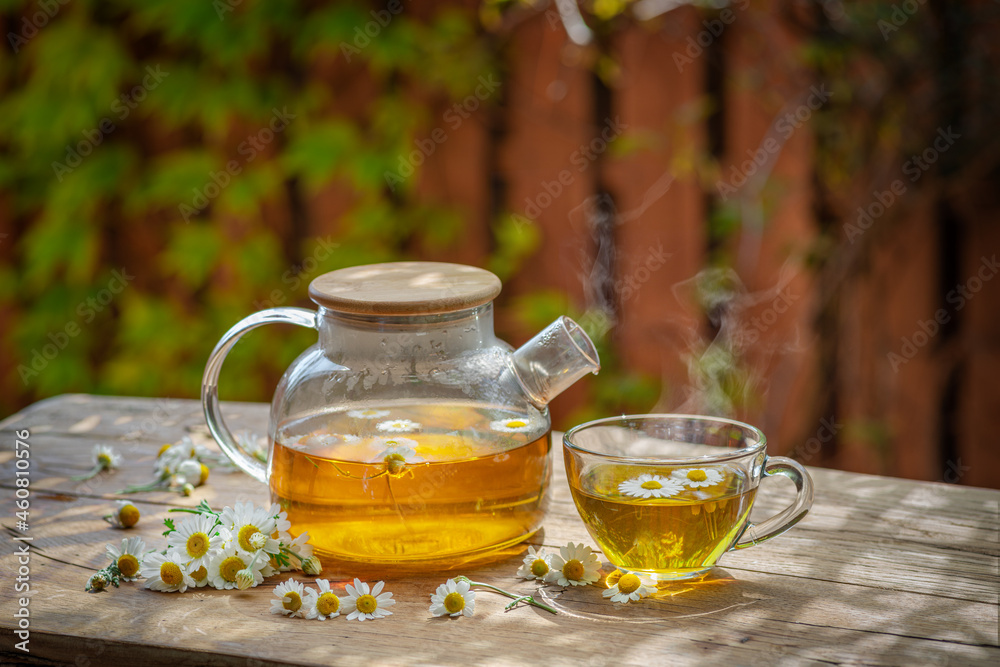 Herbal chamomile tea and chamomile flowers near teapot and tea glass on wooden table. Countryside background.
