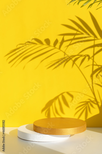 White and gold cylindrical scenes on a white table on a yellow background with a shadow of palm leaves. Premium podium for advertising product. Empty showcase for eco cosmetic product presentation.