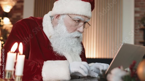 Medium close-up of white-bearded Santa Claus wearing eyeglasses, sitting at desk indoors, typing on laptop computer, then getting surprised and looking on camera photo