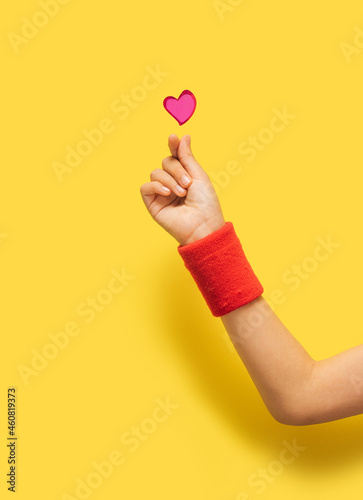 K pop concept. A girl hand with red sport cotton sweat bands showing fingers heart gesture. Red glitter heart above. Optimistic yellow in background.