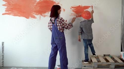A woman standing next to a child applies orange paint to the wall in her home with a paint brush with her back to the camera.