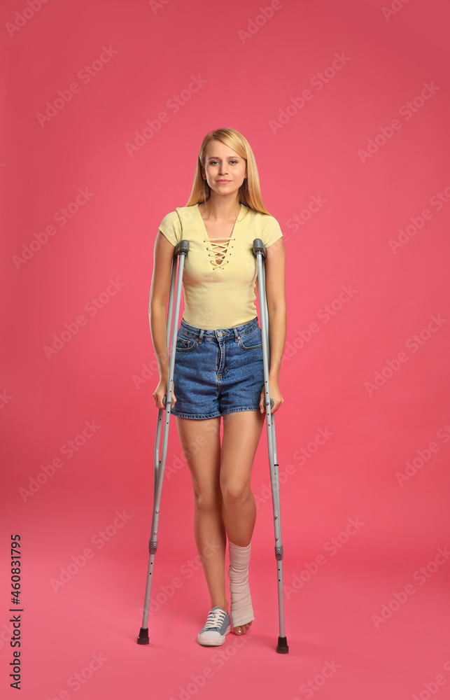 Young woman with axillary crutches on pink background