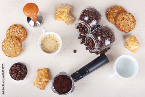 Cup of coffee, crackers, cookies, holder with ground coffee, tamper and cans of coffee beans on table. View from above.