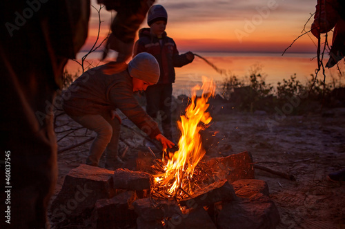 Kids of different ages sit near fire on autumn seashore after sunset, communicate and have a good time together, trust and friendship, active family weekend. Autumn outside lifestyle. Focus on fire