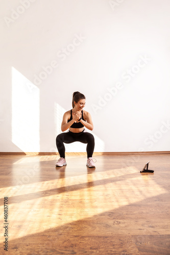 Sporty girl doing squatting sit up exercise while repeating after fitness coach online, using tablet, wearing black sports top and tights. Full length studio shot illuminated by sunlight from window.