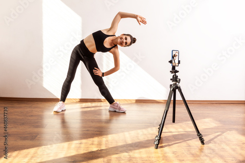 Fototapeta Naklejka Na Ścianę i Meble -  Online fitness trainer recording class of stretching, posing bending body with raised arm aside, wearing black sports top and tights. Full length studio shot illuminated by sunlight from window.
