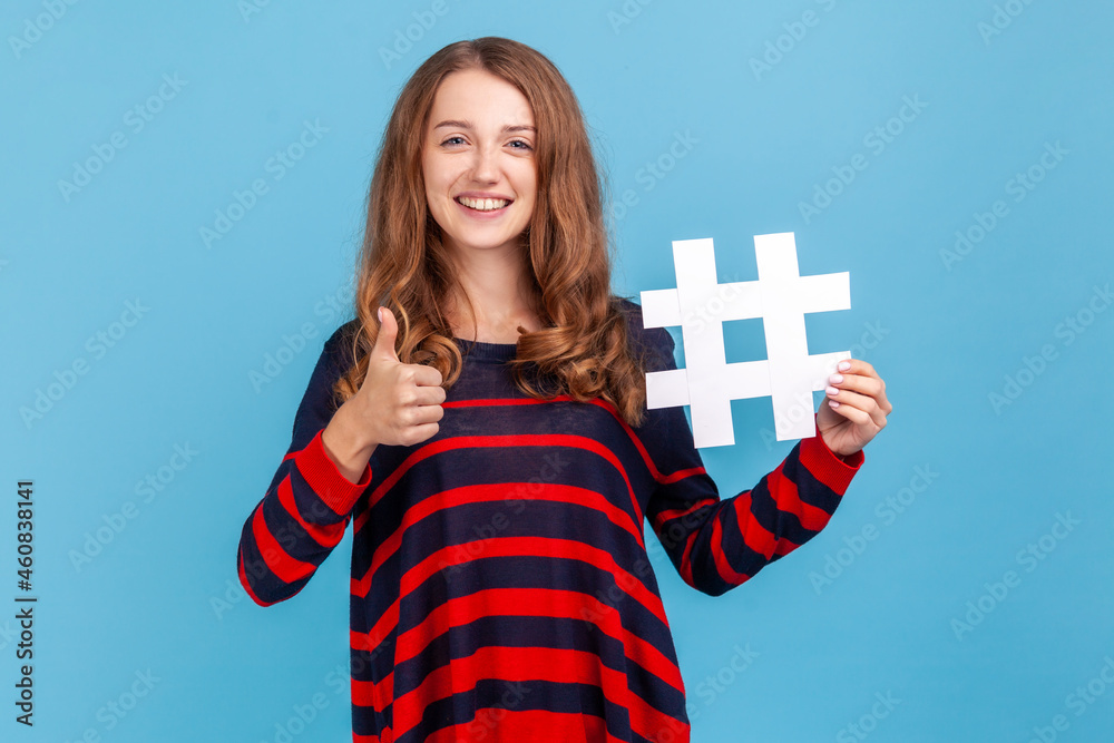 Happy woman wearing striped casual style sweater, showing hashtag and thumb up, promoting viral topic in social network, tagging blog trends. Indoor studio shot isolated on blue background. Happy woman wearing striped casual style sweater, showing hashtag and thumb up, promoting viral topic in social network, tagging blog trends. Indoor studio shot isolated on blue background.