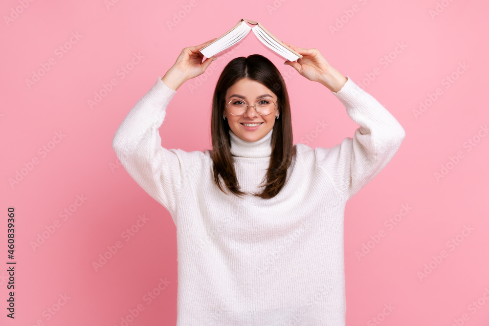 Good looking female in glasses holding opened book above head, expressing positive, education, wearing white casual style sweater. Indoor studio shot isolated on pink background.