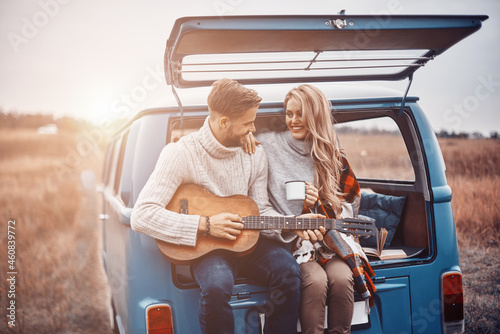 Handsome young man playing guitar for his girlfriend while both sitting in the car trunk outdoors