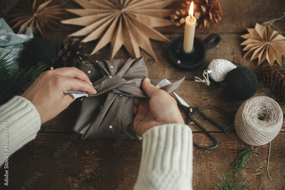 Furoshiki Christmas gift. Hands wrapping christmas gift in brown fabric on rustic wooden table with scissors, craft paper star, candle. Atmospheric moody time, nordic style. Zero waste holiday - obrazy, fototapety, plakaty 