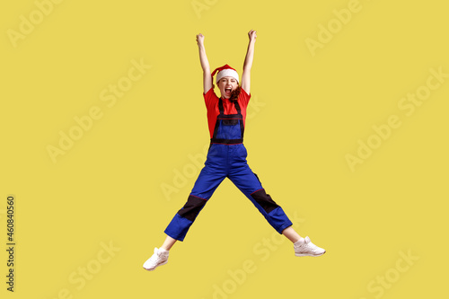 Full length portrait of excited happy female worker jumping high, finishing work before Christmas, wearing blue overalls and santa claus hat. Indoor studio shot isolated on yellow background.