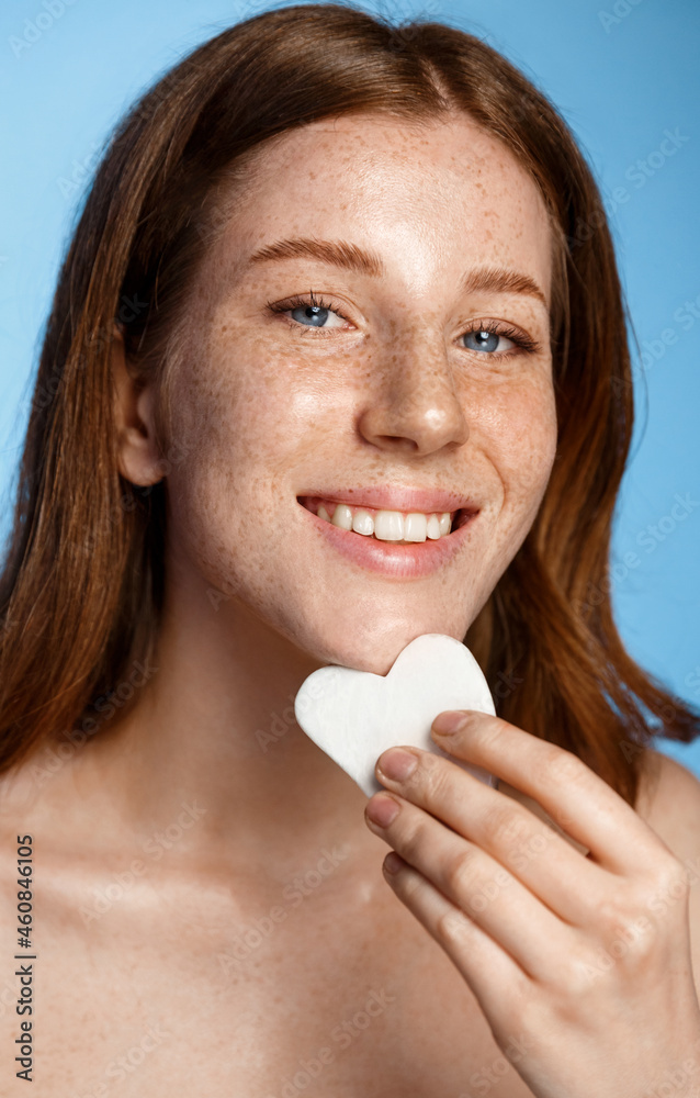 Vertical portrait of smiling redhead female model, self-massage facial skin with guasha scrapper, scrapping face and smiling, standing over blue background Vertical portrait of smiling redhead female model, self-massage facial skin with guasha scrapper, scrapping face and smiling, standing over blue background