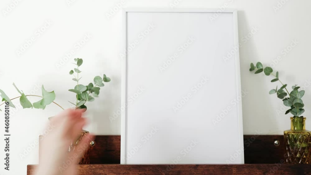 Female hand placing white picture frame on brown shelf against white wall. Mock up. Copy space. 4K UHD footage