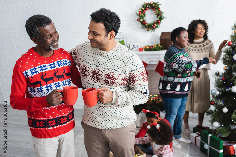 happy african american men with cups talking near blurred women and kids decorating christmas tree