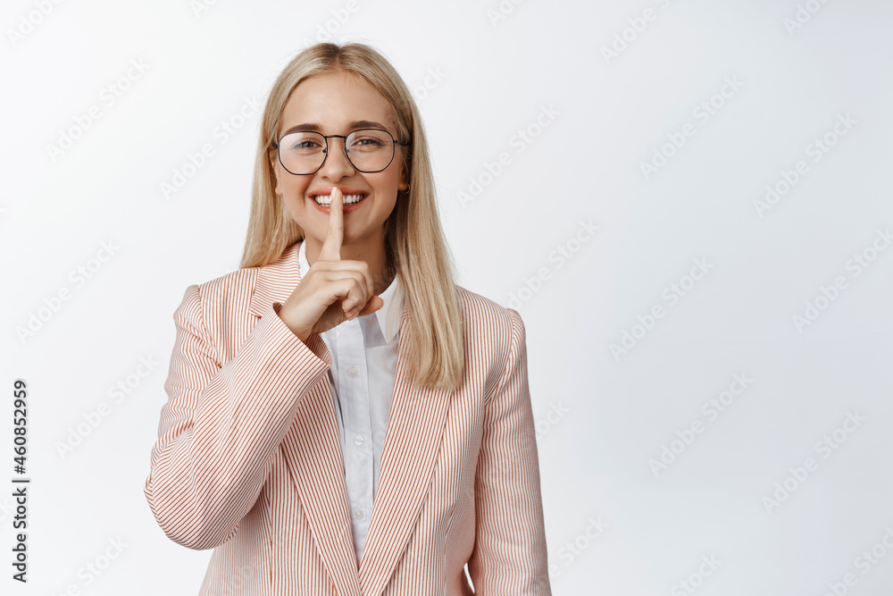 Smiling business woman showing hush sign, keeping a secret, make shh gesture, standing in suit and glasses against white background Smiling business woman showing hush sign, keeping a secret, make shh gesture, standing in suit and glasses against white background